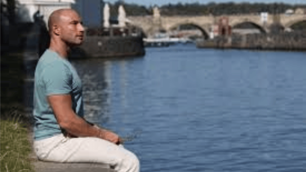 Pascal Bachmann sitting by the water, wearing a light blue t-shirt and white pants, with a historic bridge in the background.